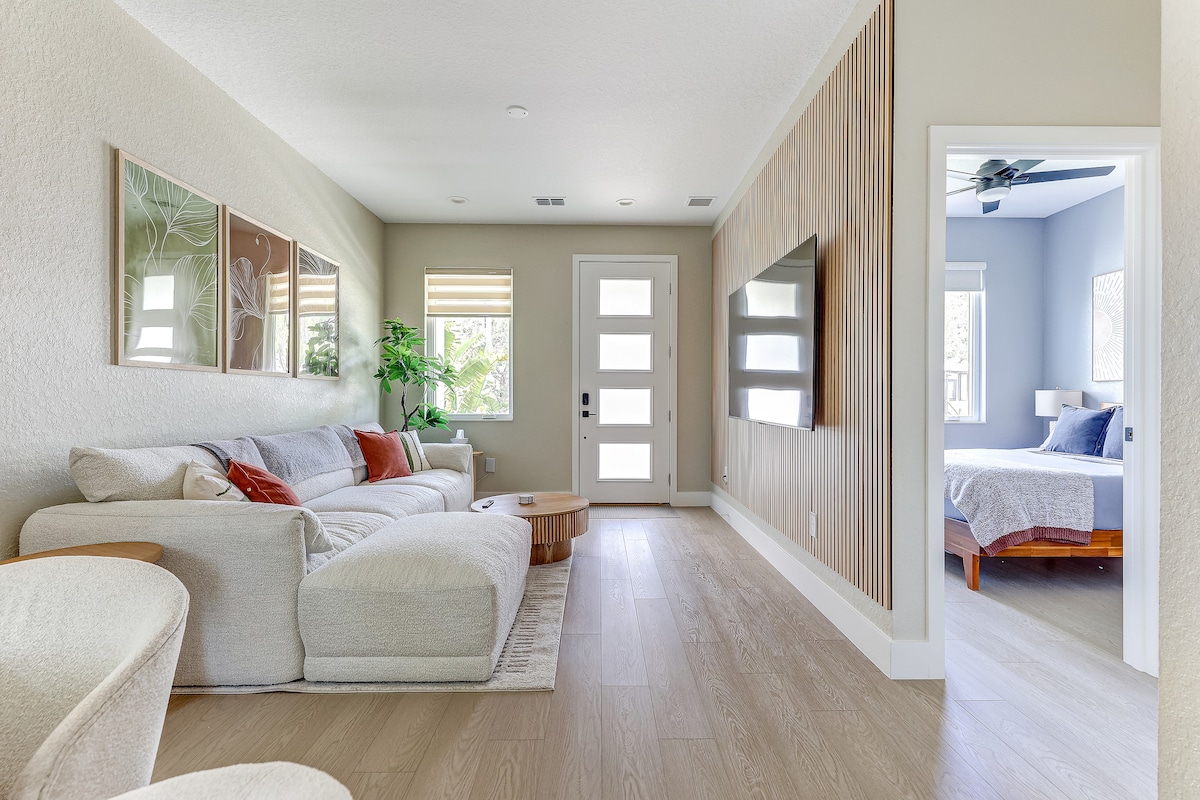 A spacious living area is present, featuring a large, comfortable sectional sofa and a circular wooden coffee table. Natural light enters through the front door and windows, while a flat-screen TV is mounted on the wall opposite. A glimpse of a bedroom can be seen through an adjacent doorway.