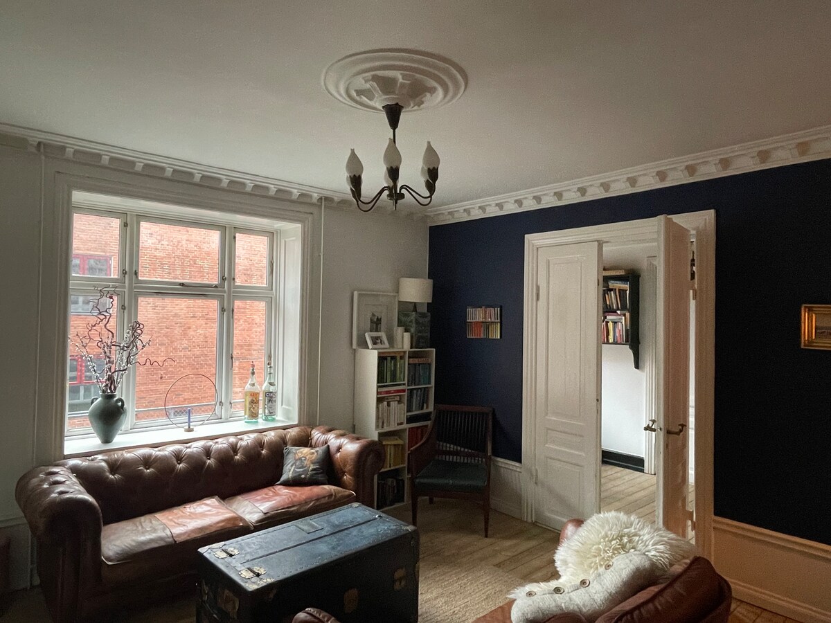 A living room is presented with large windows allowing natural light. Two leather sofas are arranged around a coffee table. A vintage trunk serves as a functional centerpiece. A bookshelf is visible, and a door leads to another room. Elegant molding enhances the ceiling.