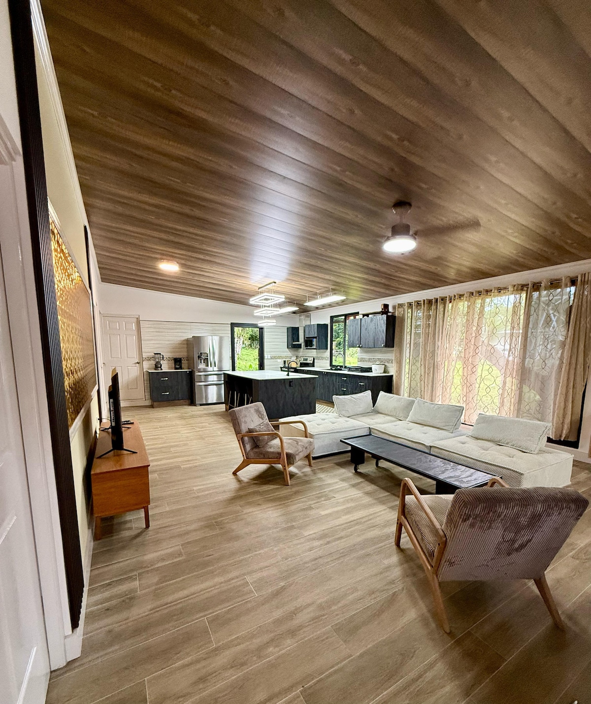 An open living area features a spacious sectional sofa and two accent chairs arranged around a low coffee table. Large windows draped with sheer curtains invite natural light, and a fully equipped kitchen with dark cabinetry can be seen in the background.