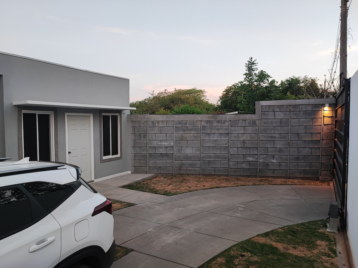 The exterior view features a sleek white vehicle parked in a gravel driveway. A linear path leads toward a modest entrance, flanked by two windows on the left. A tall, textured wall provides privacy, while greenery is visible beyond it, illuminated by soft ambient lighting.