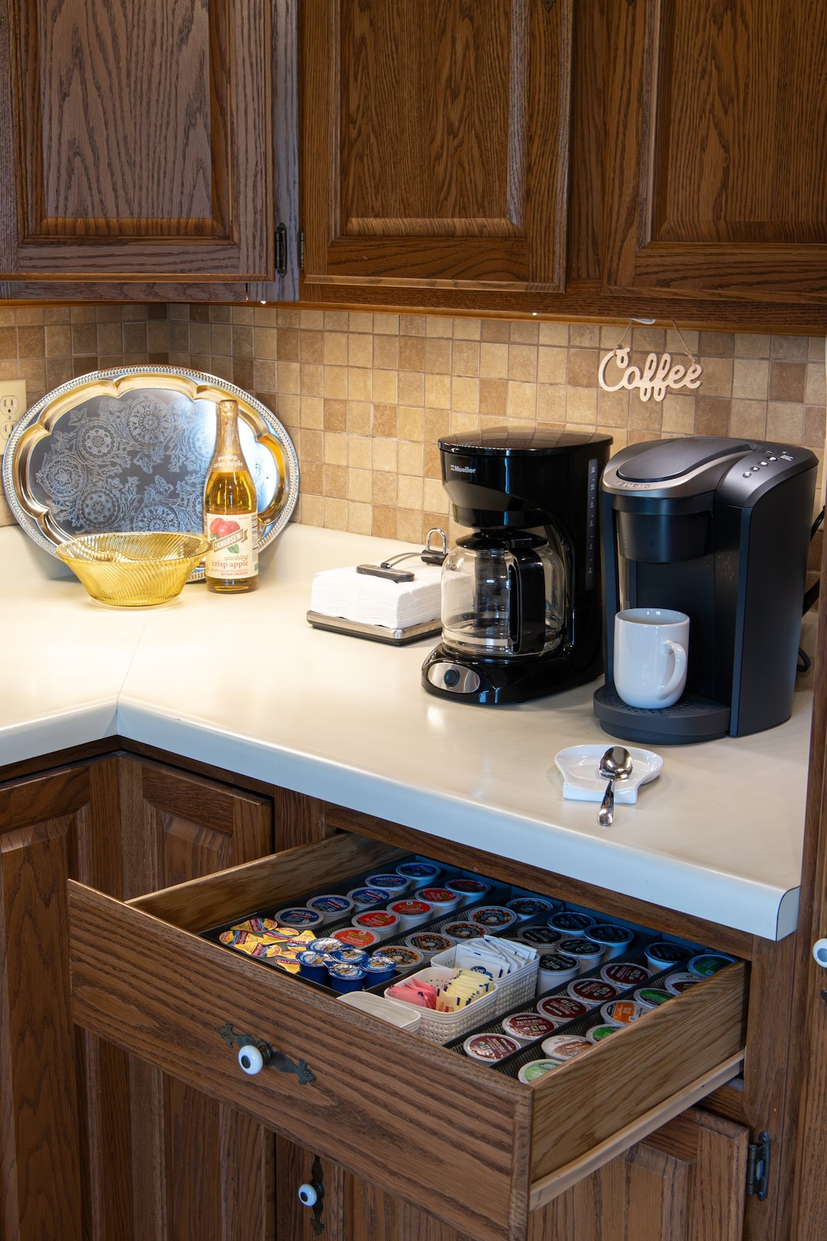 A well-equipped kitchen corner features a coffee maker, a Keurig machine, and an assortment of coffee pods neatly organized in a drawer. A decorative serving tray and sparkling beverage add a touch of charm to the countertop, surrounded by warm wooden cabinetry.