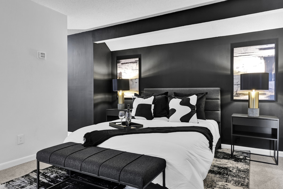 A modern bedroom showcasing a stylish black accent wall complemented by framed artwork. A spacious bed with a white comforter is highlighted by coordinating black and white pillows. Two bedside tables with lamps provide soft lighting, while a patterned rug adds texture to the floor.