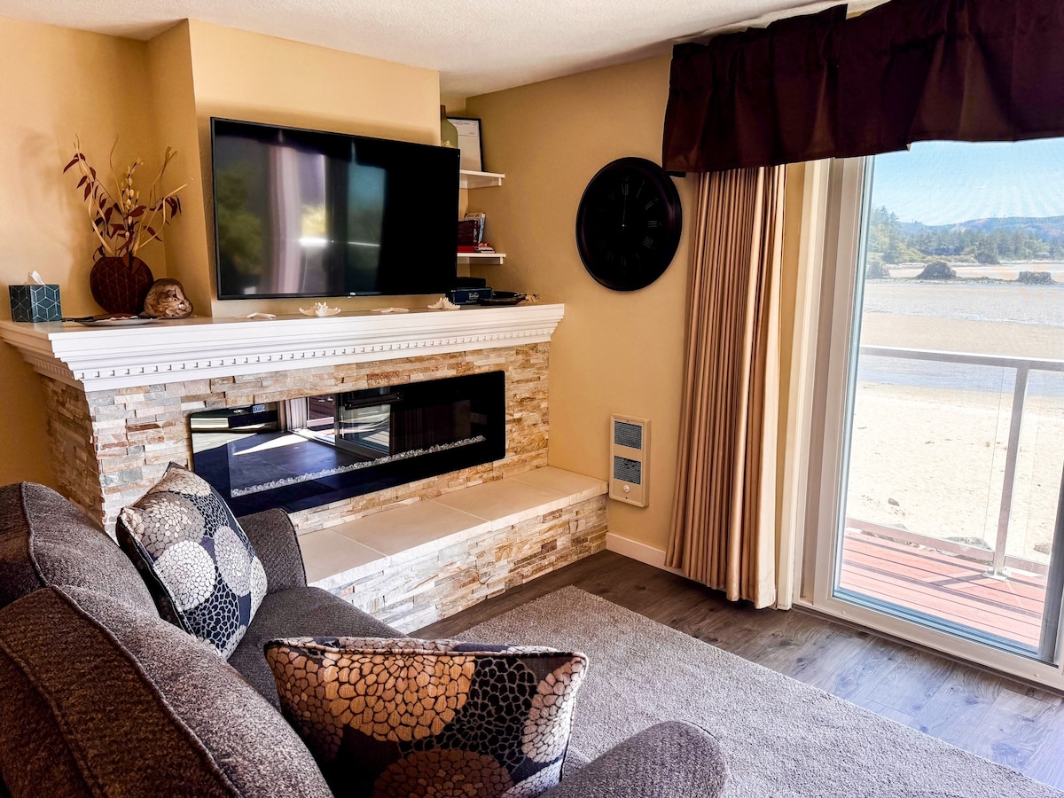 A comfortable living area is highlighted by a modern stone fireplace beneath a flat-screen television. Large windows allow natural light to fill the space, offering views of the beach and bay. Soft furnishings with textured patterns offer a cozy atmosphere.