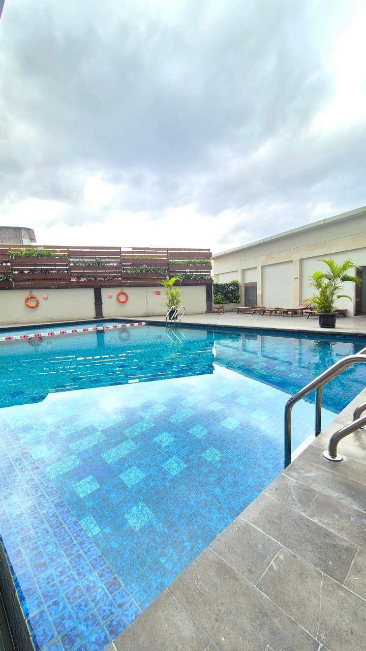 A swimming pool is showcased, featuring clear blue water and gentle ripples. Lounge chairs are arranged nearby, and palm plants provide greenery along the edges. The area is bordered by a wooden fence with lifebuoys attached, creating a serene environment for relaxation.