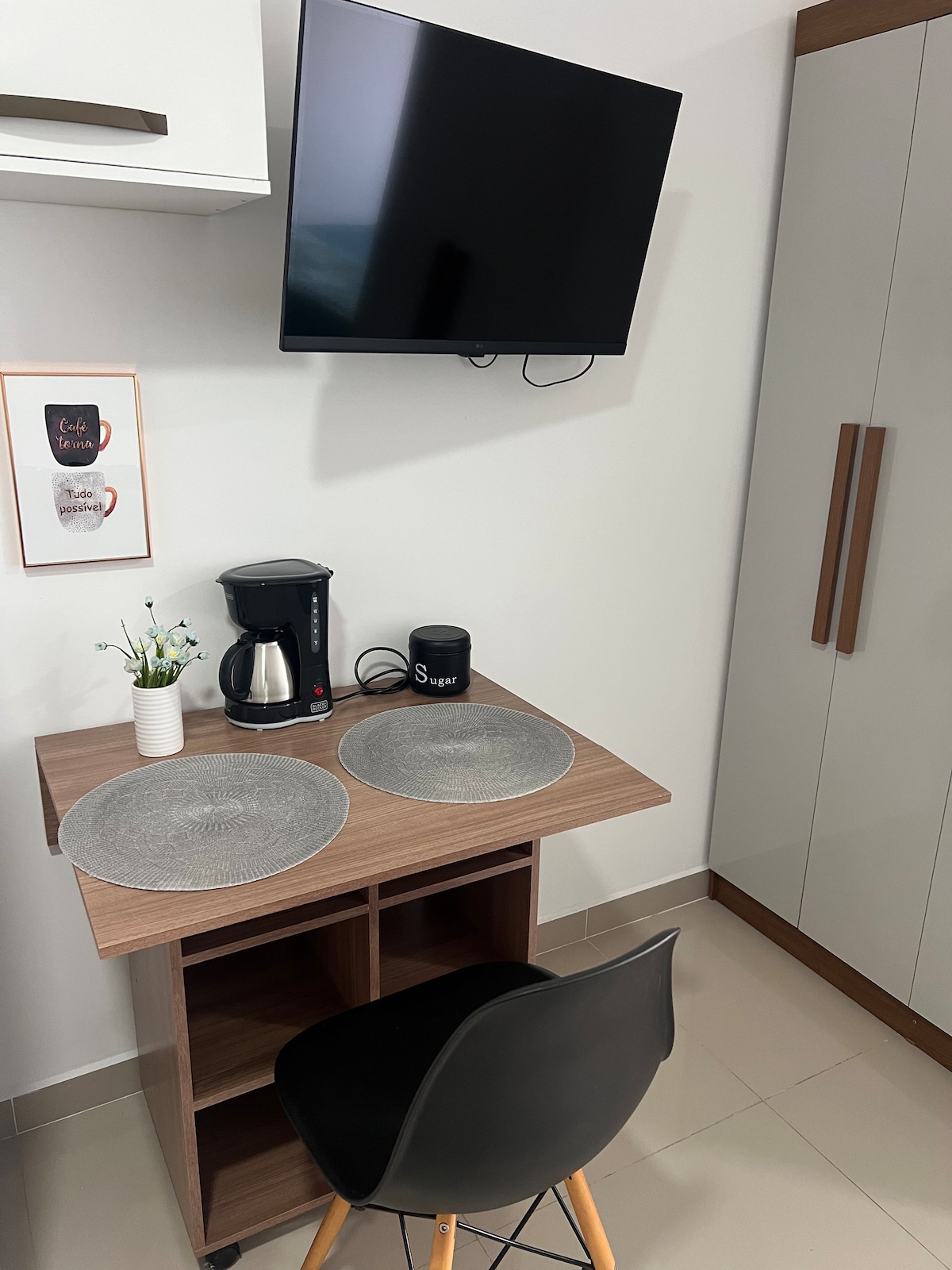 A functional dining and workspace is featured, equipped with a small wooden table and a stylish chair. A coffee maker and a coffee grinder are placed on the table, with two decorative placemats and a small potted plant. A wall-mounted television is visible above the table.
