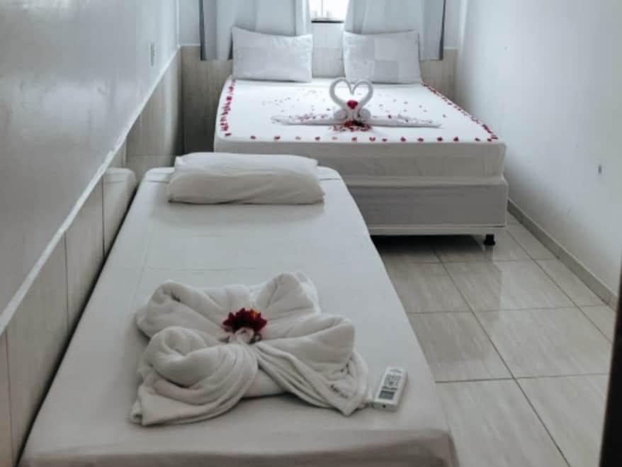 The room showcases a spacious bed adorned with decorative flower petals. Beside it, a single bed with a folded towel shaped like a swan is displayed. Soft natural light filters through curtains, illuminating the clean and minimalist design.