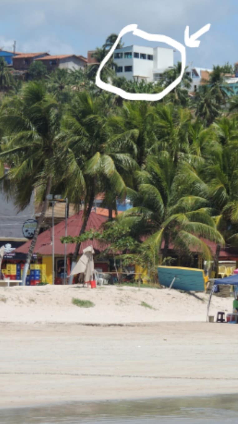 The image shows a beach scene with golden sand and palm trees in the foreground. A building is visible perched on a hill in the background, providing a view of the coastline and the surrounding area.