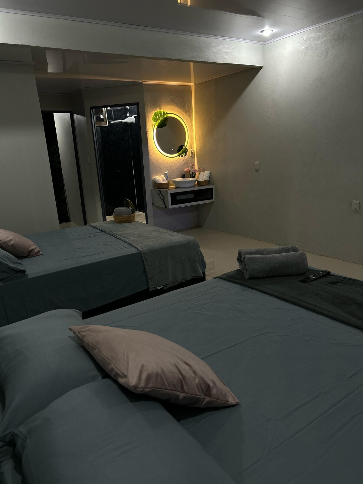 A calm and elegant space features two spacious beds covered in soft blue linens, accented with light pink pillows. A round mirror with a warm glow is positioned above a modern vanity. The bathroom area is partially visible, along with a potted plant and towels.