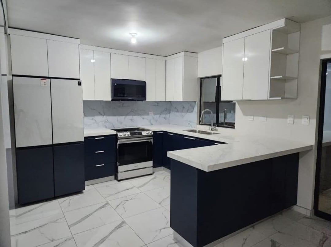 A modern kitchen is equipped with sleek cabinetry in white and navy blue. A stainless steel refrigerator and stove complement the marble-effect countertops. Ample natural light filters in through the large window, illuminating the spacious layout and clean lines of the room.