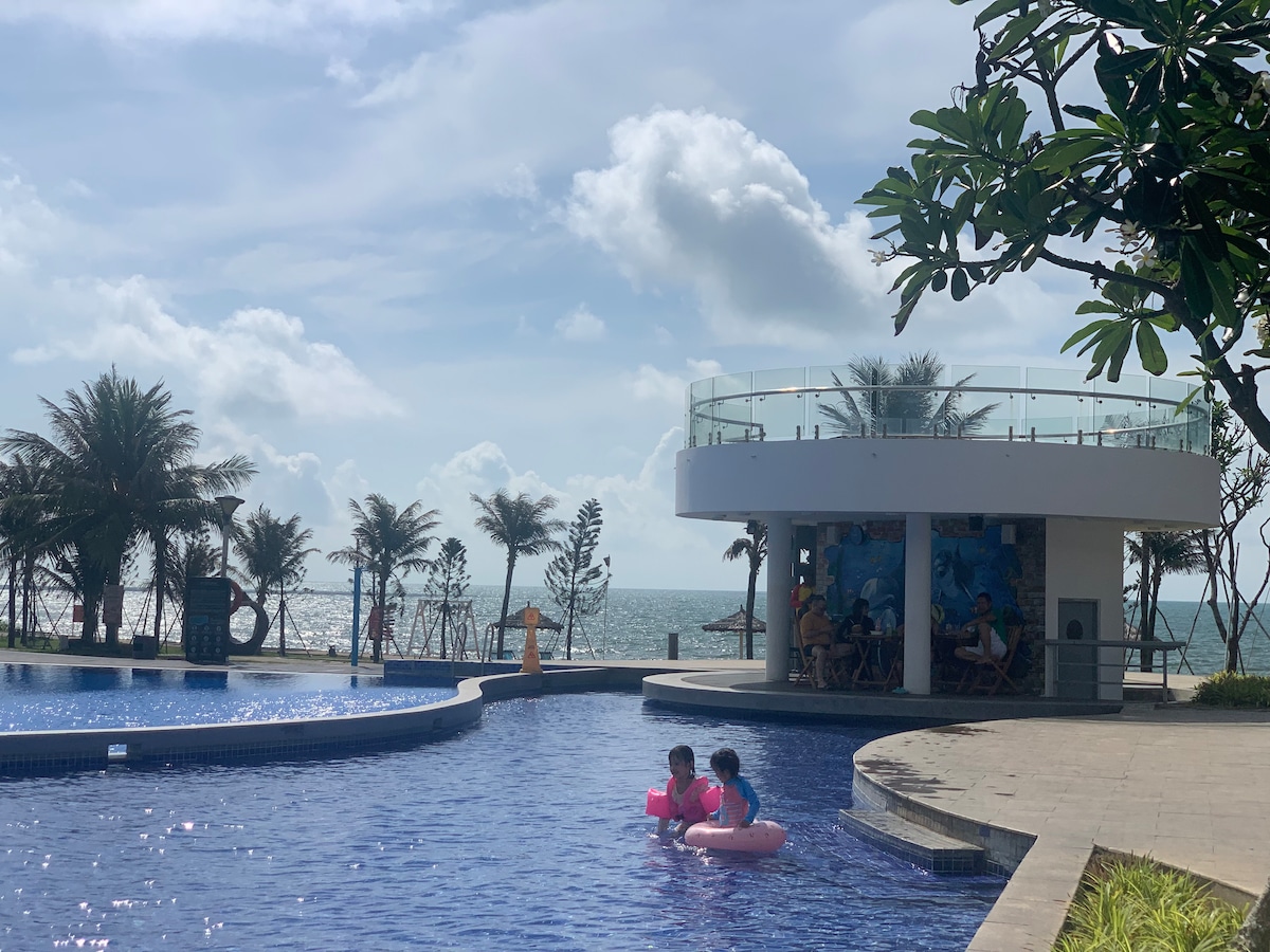 A tranquil pool area features clear blue water with gentle curves leading towards the beach. Palm trees are visible in the background, framing the scene, while a modern structure with a glass railing offers a relaxing spot for guests. The ocean extends beyond, blending with the horizon.