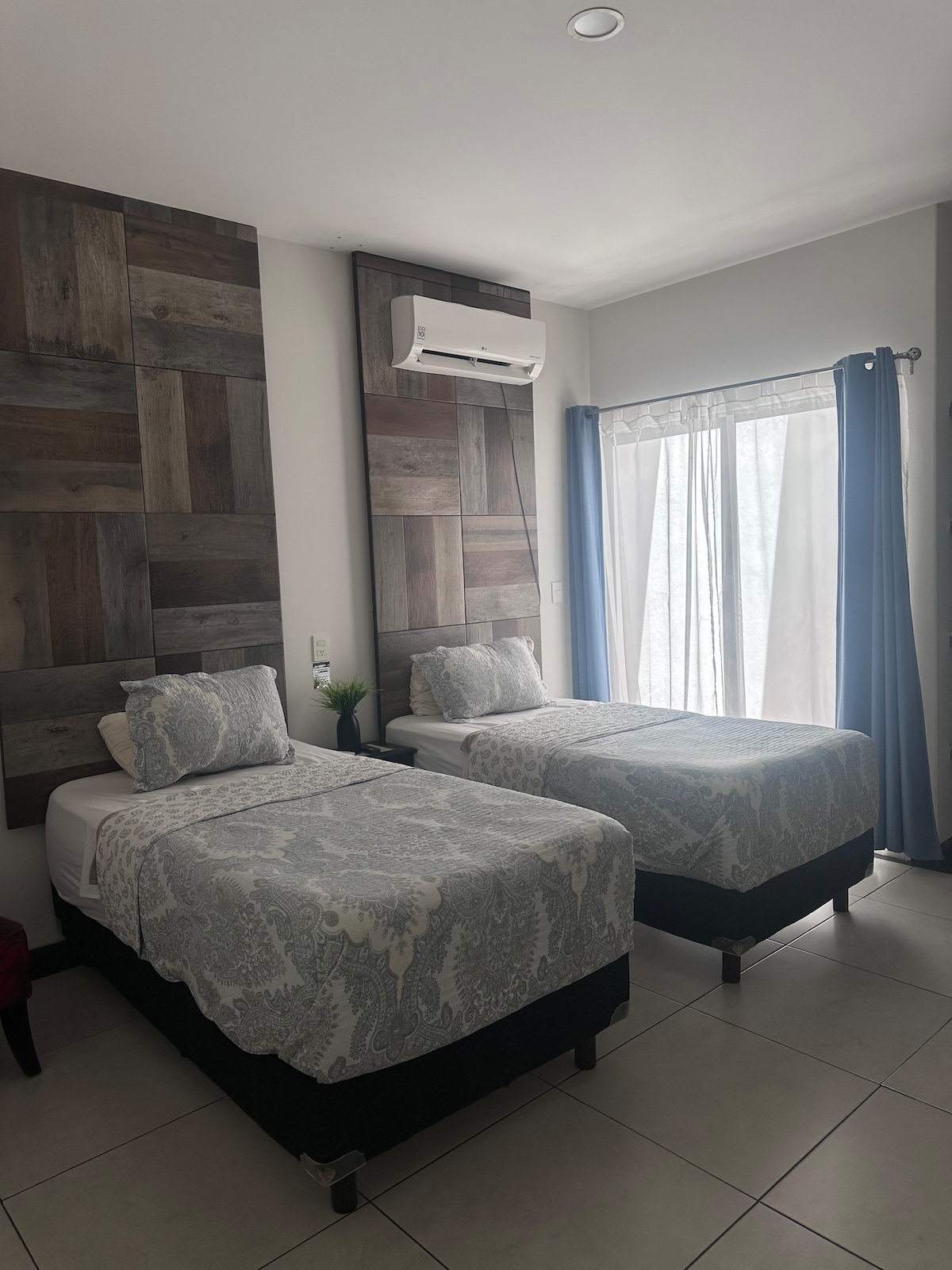 A well-lit room features two twin beds covered with patterned bedding. Light wooden paneling adorns the walls, while a large window with sheer curtains allows natural light to enter. A small plant is positioned on a bedside table, complementing the room's decor.