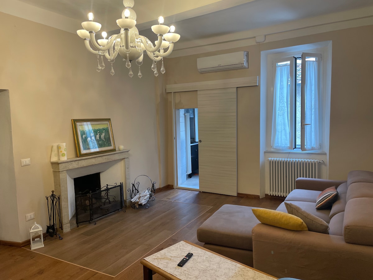The living area features a comfortable sofa, a coffee table, and an antique fireplace, enhancing the space's elegance. Natural light enters through a window with sheer curtains, and a decorative chandelier hangs from the ceiling, adding charm to the room.