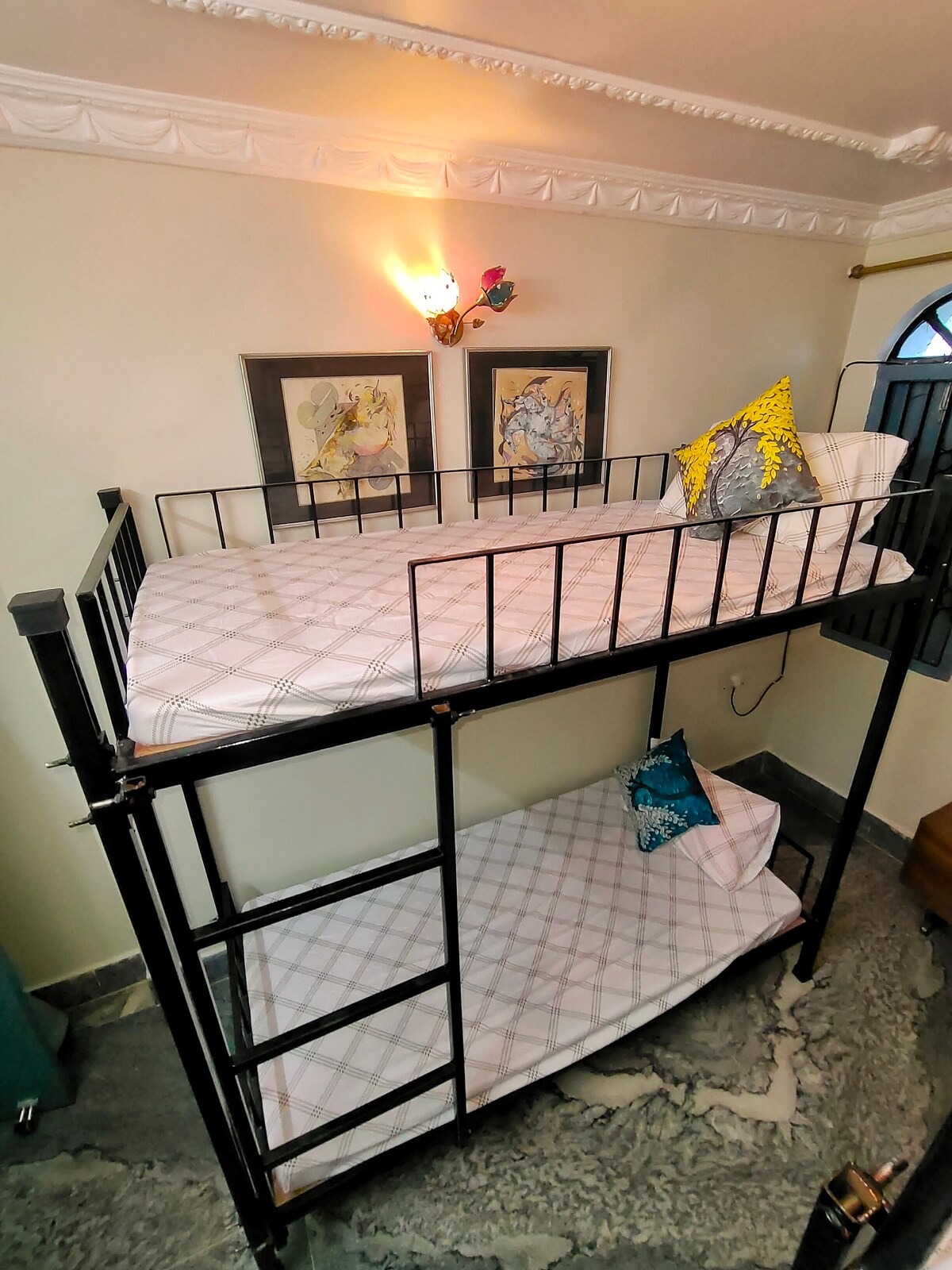 A metal bunk bed is displayed with a lightly patterned top mattress and a solid bottom mattress. Decorative pillows in vibrant colors add a touch of warmth. The room features framed artwork on the wall, and soft lighting creates a cozy ambiance.