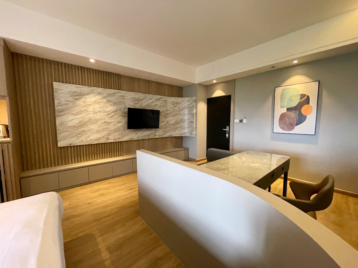 A modern studio apartment features a smooth, textured wall housing a flat-screen TV. Natural light filters through, illuminating a spacious work desk with a stone top and ergonomic chair. A minimalist design is complemented by soft wood tones and contemporary artwork on the wall.