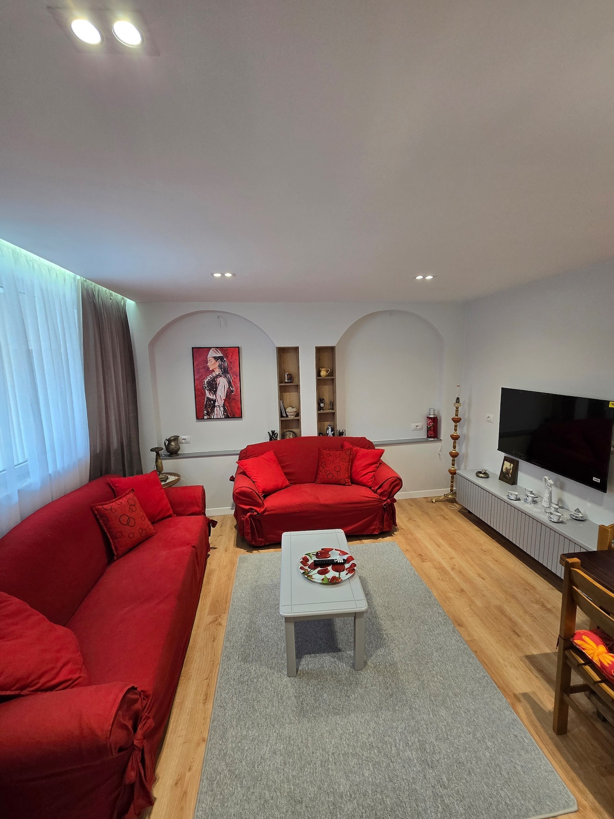A vibrant living area features two red armchairs flanking a neutral-toned sofa, complemented by a light gray area rug. A modern television is mounted on the wall, while decorative shelves hold accent pieces. Natural light filters through sheer curtains, enhancing the room's brightness.