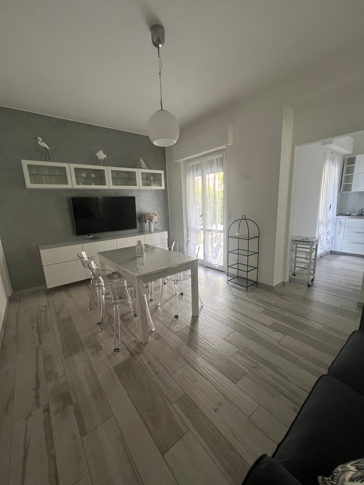 A bright living area features a sleek white table surrounded by transparent chairs. A wall-mounted television is visible above a minimalist white cabinet. Light filters through the sheer curtains, enhancing the calm atmosphere. An adjoining kitchen area is partially visible in the background.
