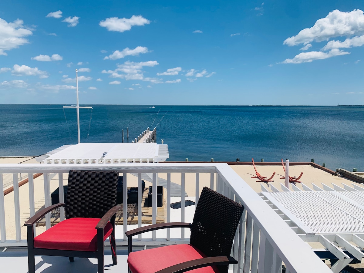 A view from a balcony is presented, featuring two red chairs positioned beside a railing. The expansive bay stretches out in the background, framed by clear blue skies and scattered clouds. A pier extends into the water, inviting relaxation and outdoor activities.
