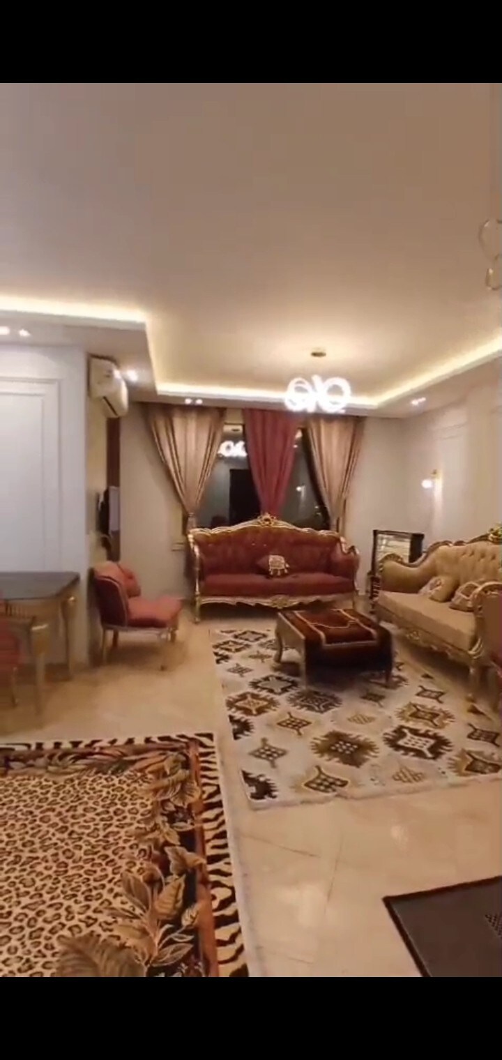 A spacious living area is adorned with elegant furniture, featuring a patterned area rug and rich curtains framing a window. The warm lighting is complemented by decorative light fixtures overhead, creating a comfortable gathering space with plush seating arrangements.