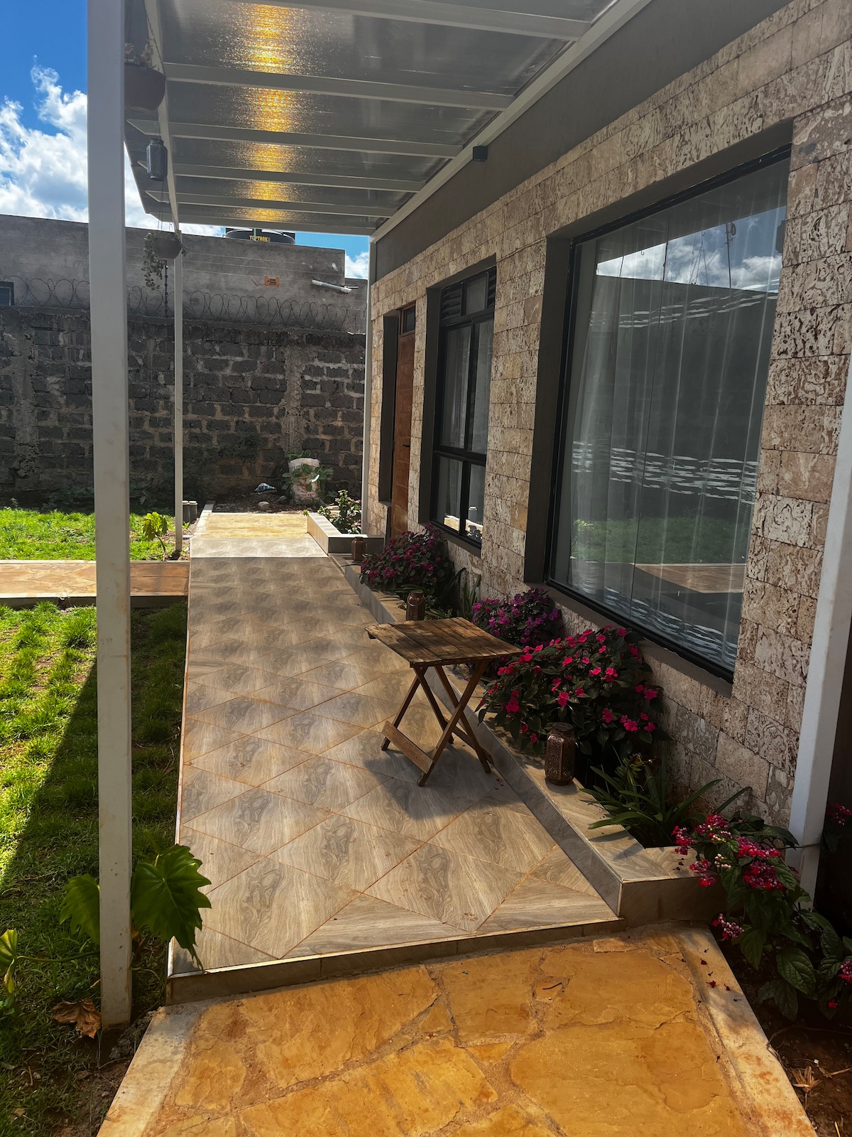 A charming pathway lined with colorful flowers leads to a spacious entrance. The stone facade of the building adds texture, while a small wooden chair is placed nearby. Natural light casts gentle shadows on the patterned tiles, providing a welcoming outdoor space.