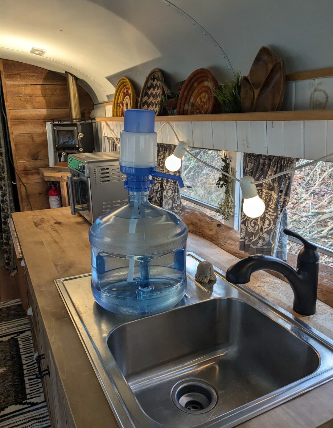 A modern kitchenette is presented with a stainless steel sink and a countertop featuring a large water dispenser. Under-cabinet lighting illuminates the space, while natural wood accents are visible on the cabinets and decor. Decorative items are arranged on shelves above the counter.