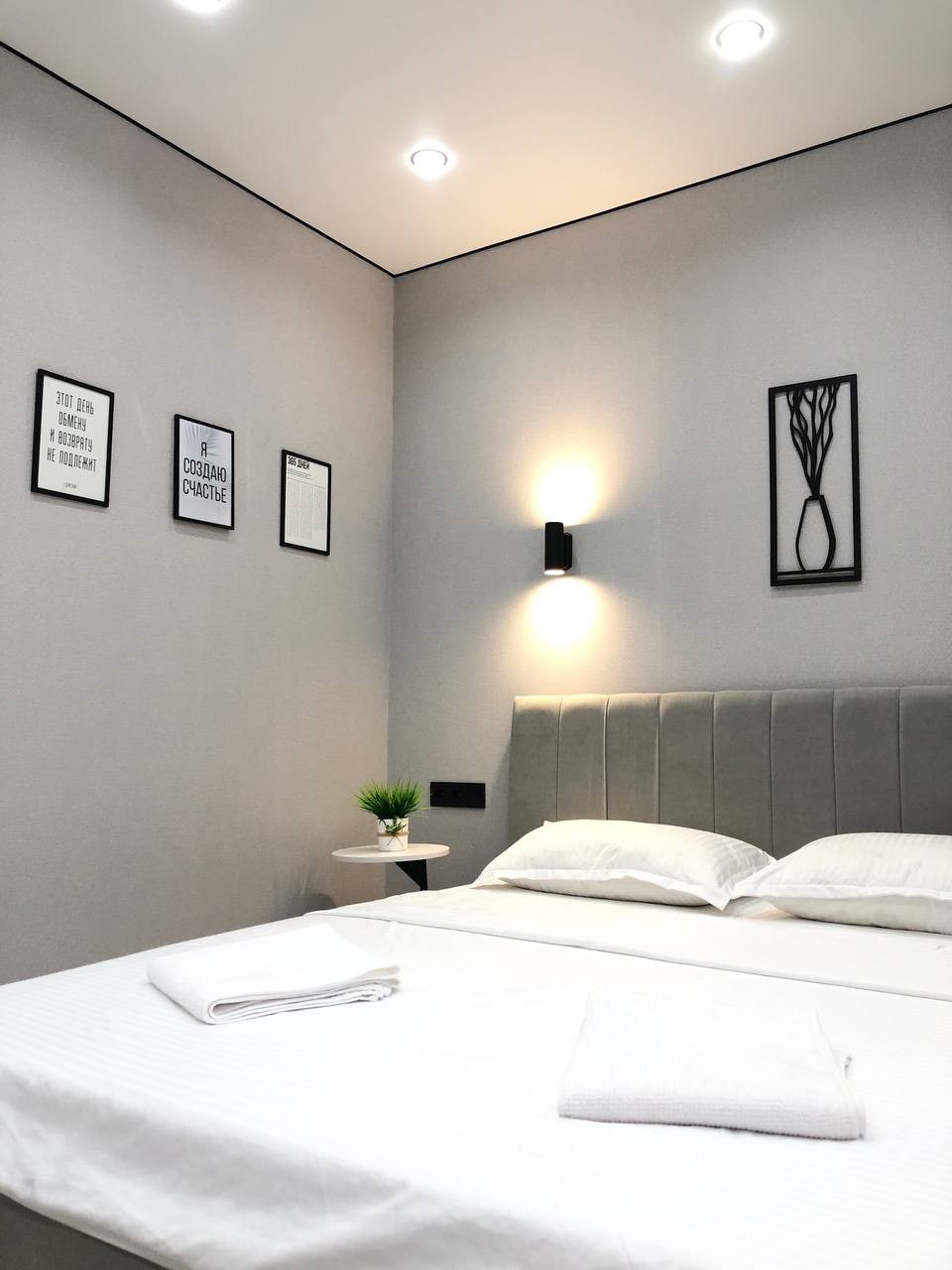 A modern bedroom features a comfortable bed with fresh linens and neatly arranged towels. Minimalist wall art complements the simple decor. Soft lighting from wall sconces enhances the ambiance, while a small potted plant adds a touch of greenery to the space.