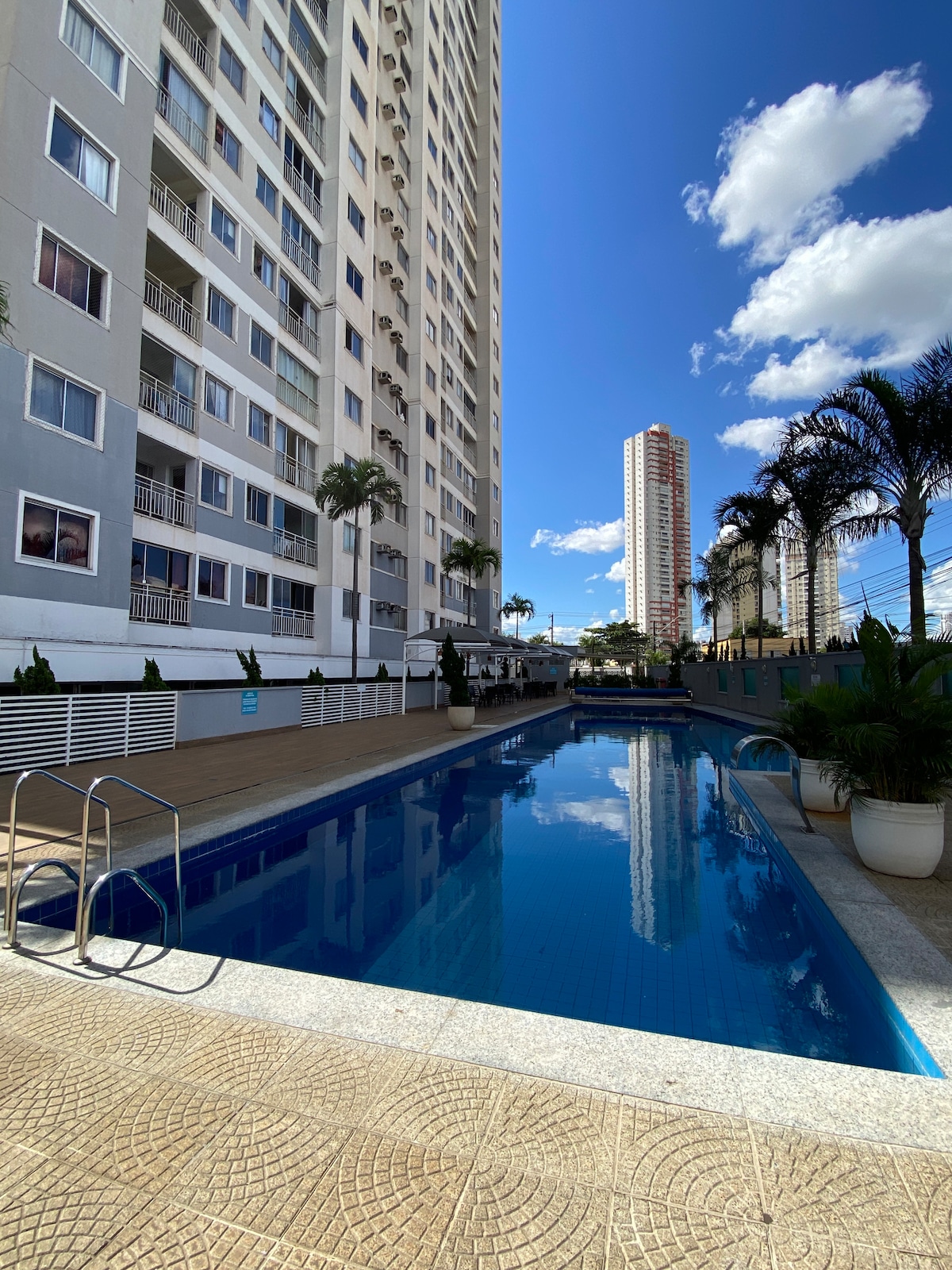 A tranquil pool area is bordered by a clean deck, with palm trees providing a natural touch. The modern building looms in the background, reflecting a bright blue sky dotted with clouds. Lounge chairs line the edge of the pool, inviting relaxation.