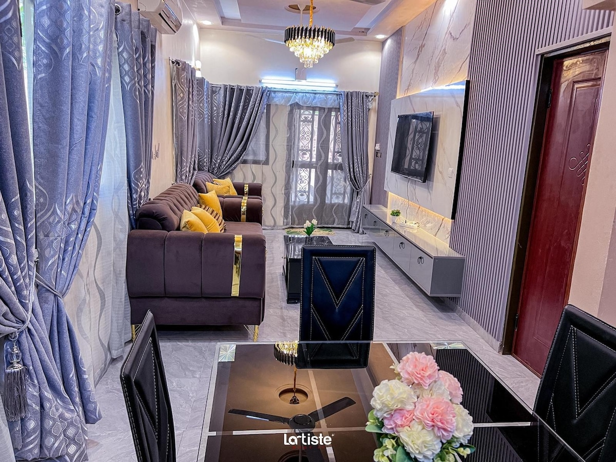 The living area features a comfortable sofa accented with gold detailing, arranged around a stylish glass coffee table. Large windows allow natural light to fill the space, while elegant curtains frame the view. A sleek entertainment unit is mounted on the wall, displaying a television.