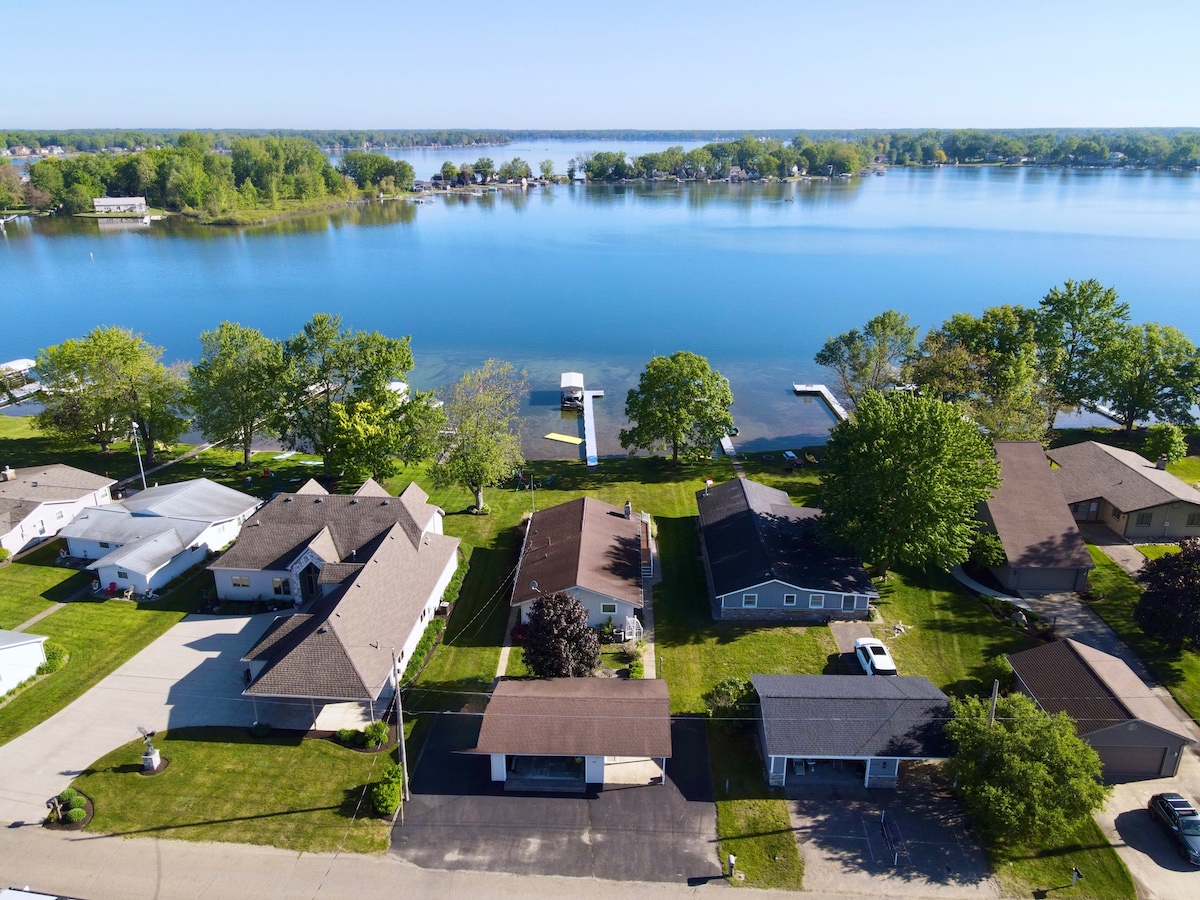 An aerial view captures the serene Coldwater Lake, showcasing the clear blue water surrounded by lush greenery. Several cottages are visible along the shoreline, accompanied by private docks extending into the lake, while the surrounding area features manicured lawns and tree-lined streets.
