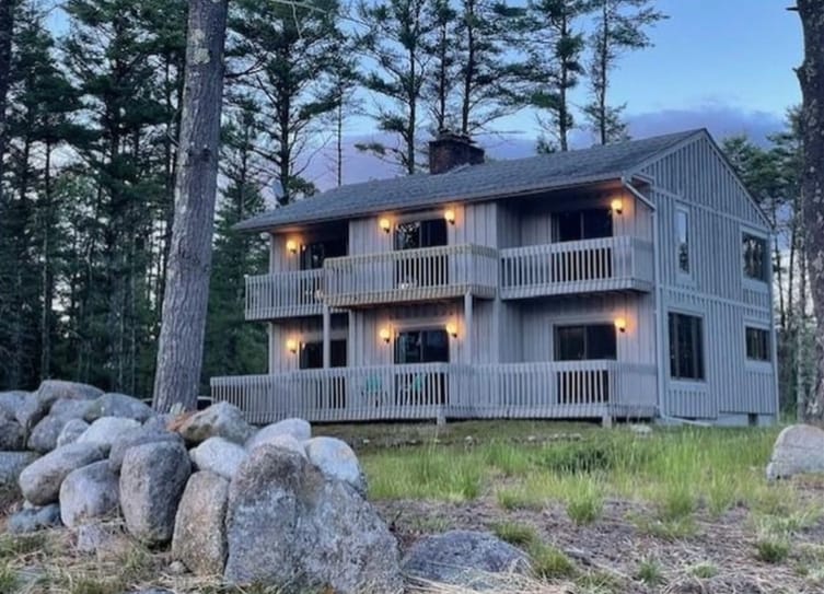 A two-story lake house is surrounded by tall trees, showcasing wooden balconies on both levels. The exterior is painted in a light color, with warm lights illuminating the space. A rocky area in the foreground adds to the rustic charm of the peaceful environment.