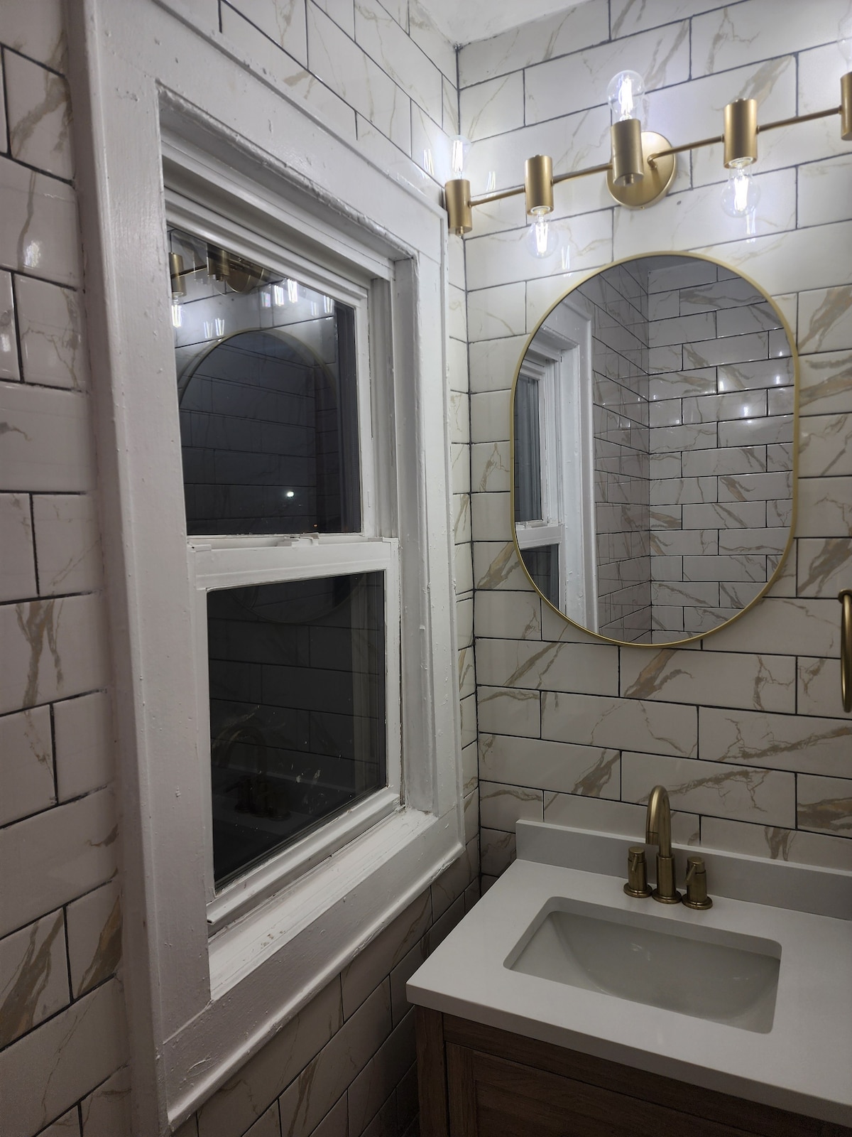 An elegant bathroom is featured with a large oval mirror above a modern sink. The walls are adorned with white and gold veined tiles, complemented by a window that allows natural light to enter. Stylish light fixtures enhance the ambiance.
