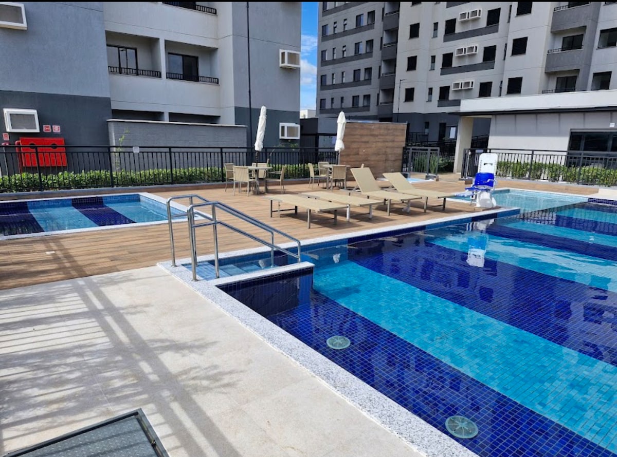 A modern swimming pool area is visible, featuring multiple pools with clear blue water. Sun loungers are arranged around the pool deck, which is complemented by a wooden area for relaxation. Additionally, lawn space is seen beside the pools, enhancing the open atmosphere.