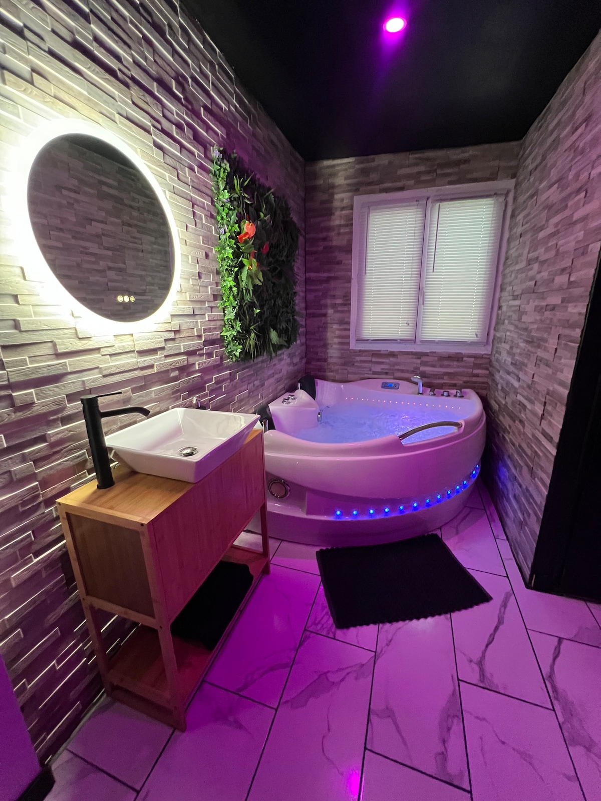 A spa area features a circular mirror illuminated with soft light above a wooden vanity. A whirlpool bath with ambient lighting is positioned nearby, surrounded by textured stone walls. A window partially covered by white blinds allows natural light to enter, enhancing the soothing space.