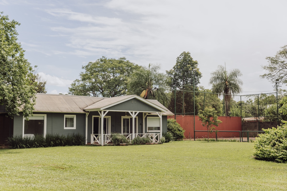 A spacious exterior offers a view of a traditional family villa surrounded by lush greenery. The building features a covered porch and is adjacent to a tennis court. The well-maintained lawn enhances the natural setting.