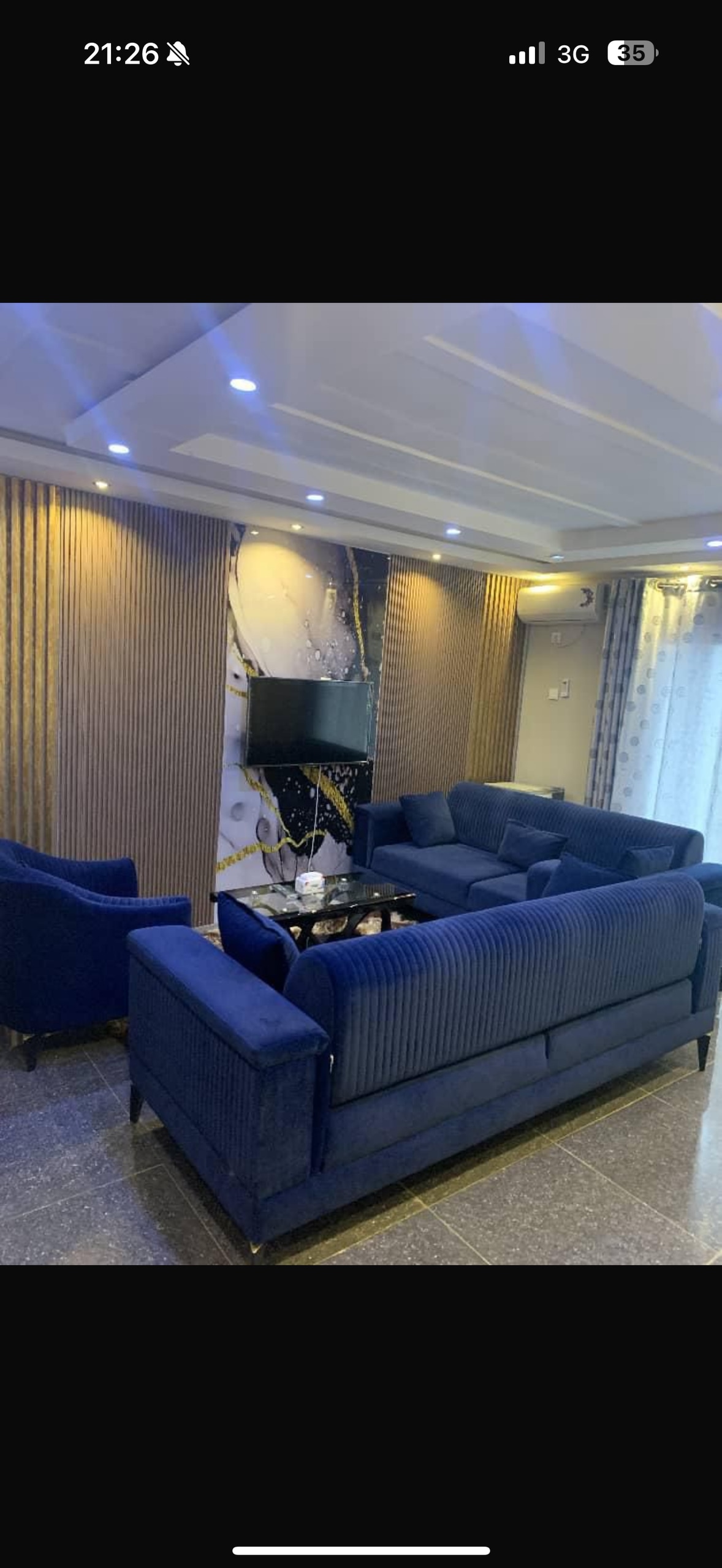 A stylish living area is furnished with a blue sofa set and matching armchairs. A coffee table is positioned at the center, complemented by decorative items. Soft lighting and elegant wall coverings enhance the serene environment, while a wall-mounted television is visible.