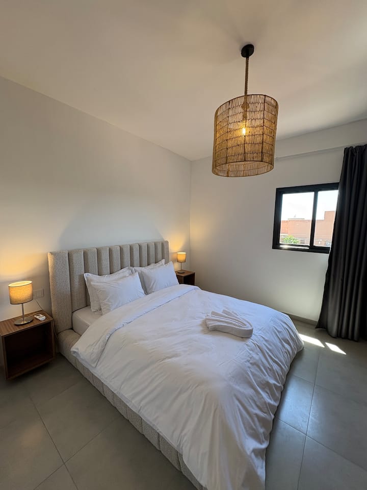 Un sexto dormitorio elegante con una gran cama de matrimonio, una cabecera acolchada gris y una lámpara de mimbre trenzado. Luminosa y con aire acondicionado, ofrece un entorno sereno para descansar después de un almuerzo o una cena en familia.