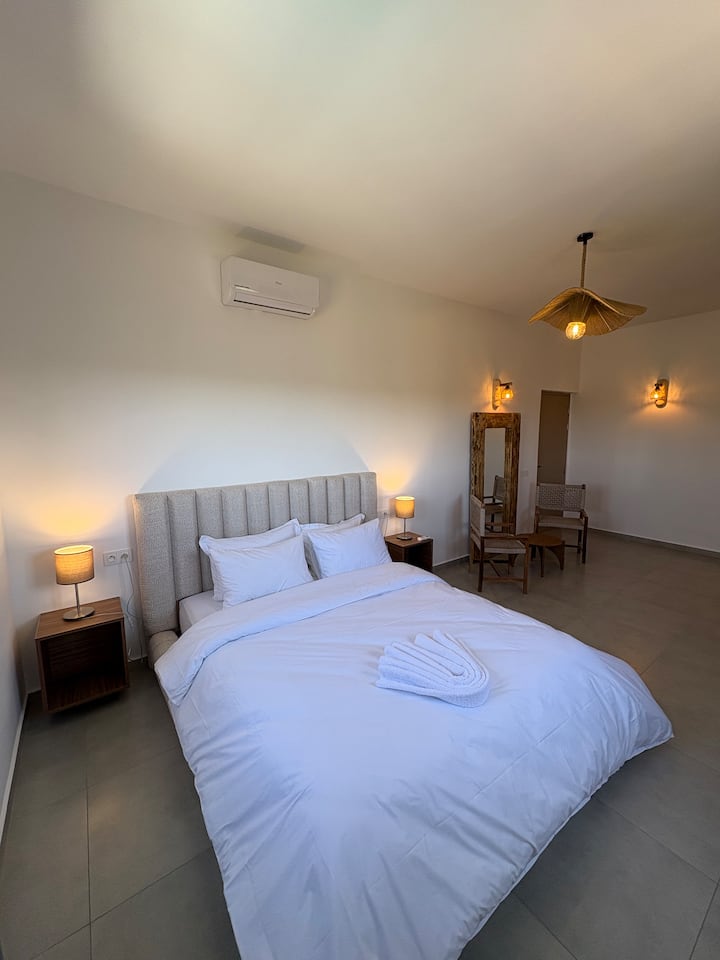Amplia suite con aire acondicionado y una elegante cama de matrimonio, una zona de estar privada con sillones de madera y un gran espejo rústico. La habitación está sublimada por una lámpara de diseño de mimbre y lámparas de apoyo que crean un ambiente suave.