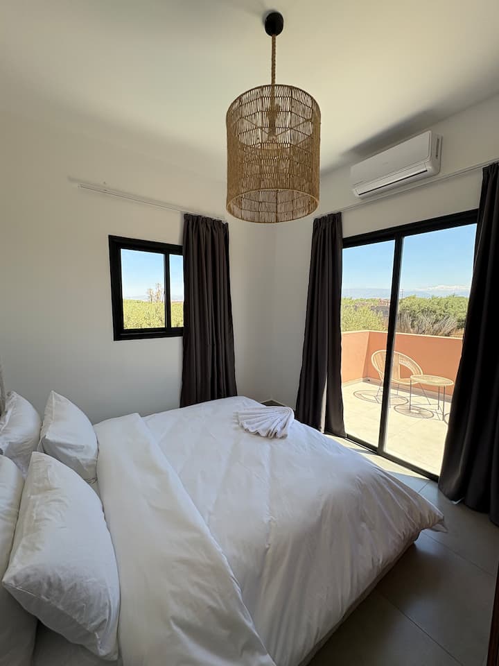 Tercer dormitorio luminoso con una espaciosa cama de matrimonio y decoración natural. Se abre a un balcón privado con muebles de exterior, ideal para relajarse. La habitación está equipada con una lámpara de araña de mimbre y aire acondicionado.
