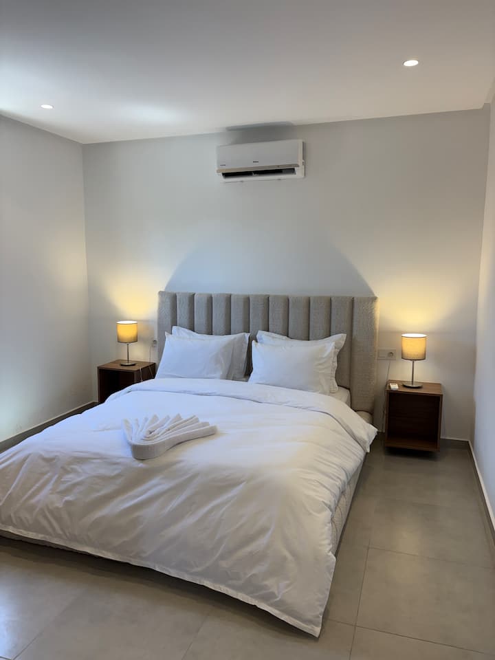 Habitación principal serena con una gran cama doble, una cabecera acolchada gris y ropa de cama blanca impecable. Incluye mesitas de noche de madera con lámparas de apoyo, aire acondicionado e iluminación suave.