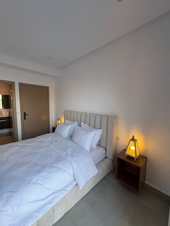Segundo dormitorio elegante con una cómoda cama de matrimonio, una cabecera de terciopelo gris y lámparas de cabecera artesanales de ratán. La habitación tiene aire acondicionado y acceso directo a un moderno baño privado.