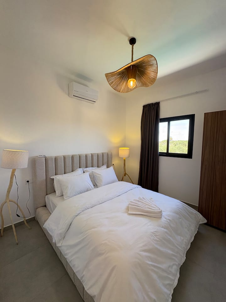 El cuarto dormitorio ofrece un diseño artístico con una lámpara de araña de mimbre tallada y lámparas de pie de madera flotante. Cuenta con una acogedora cama doble, aire acondicionado y una ventana que se abre a la naturaleza circundante.