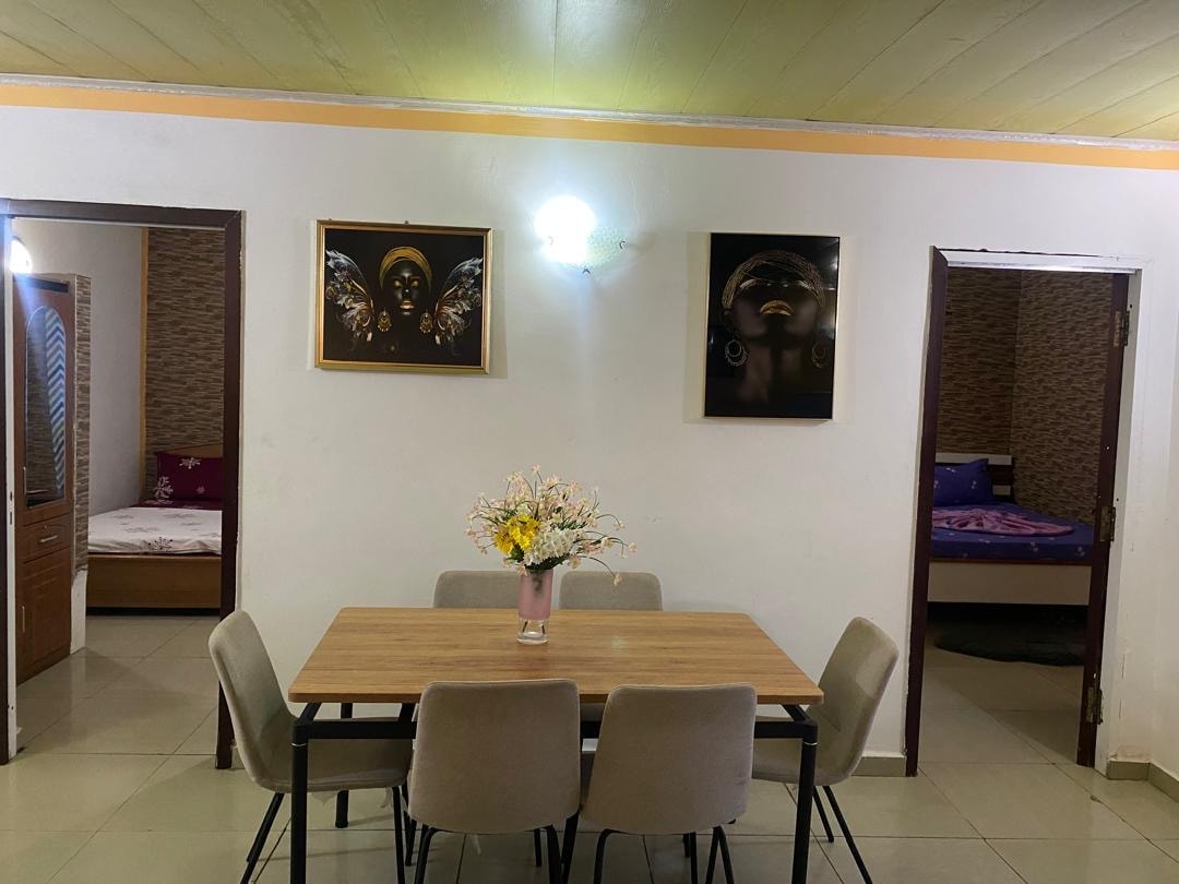 A dining area features a rectangular table surrounded by four upholstered chairs. On the wall, two framed artworks add visual interest. Open doorways lead to adjoining sleeping areas, each displaying cozy bedding against neutral walls.