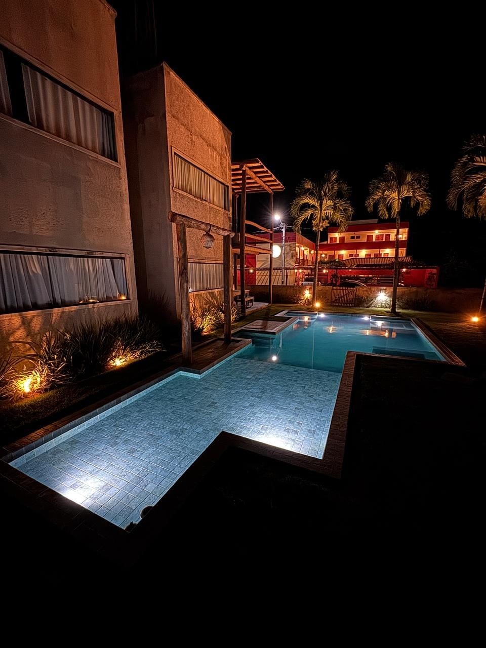 The exterior highlights a pool area illuminated by soft lighting at night. The water reflects the warm glow, while palm trees are visible in the background. Nearby buildings are subtly lit, enhancing the calming ambiance of the outdoor space.