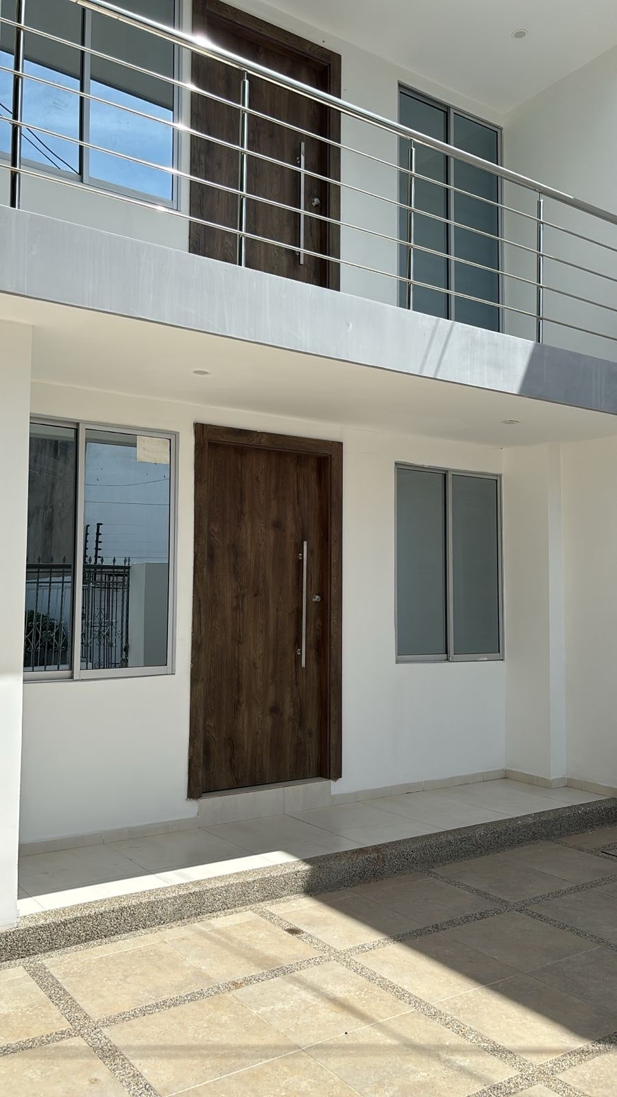 The exterior of the apartment is highlighted by a sleek wooden door and modern railings. Large windows provide natural light and a view of the entrance space, which features a clean, tiled floor and concrete steps leading to the entrance.