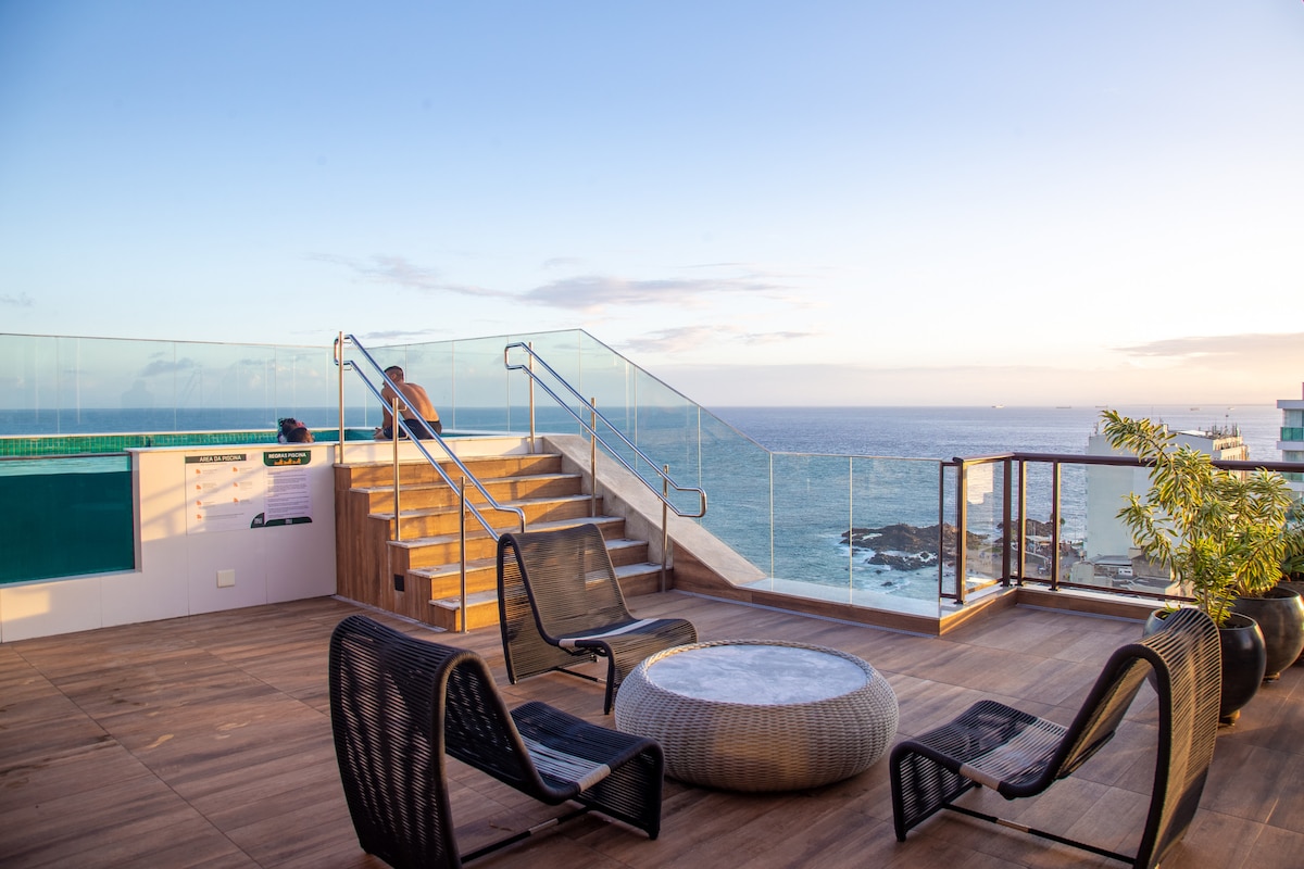The rooftop area features a seating arrangement with four woven chairs surrounding a circular table. A staircase leads down to another level, and panoramic views of the ocean are visible through the glass railing.