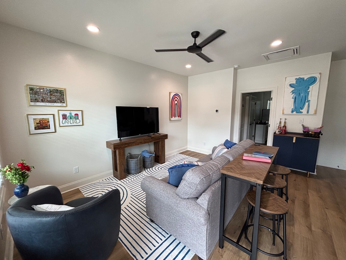A modern living area features a television mounted on the wall, complemented by a wooden console. A gray sofa is paired with a striped area rug. A small dining table with chairs occupies one corner, alongside a cozy armchair and decorative artwork on the walls.
