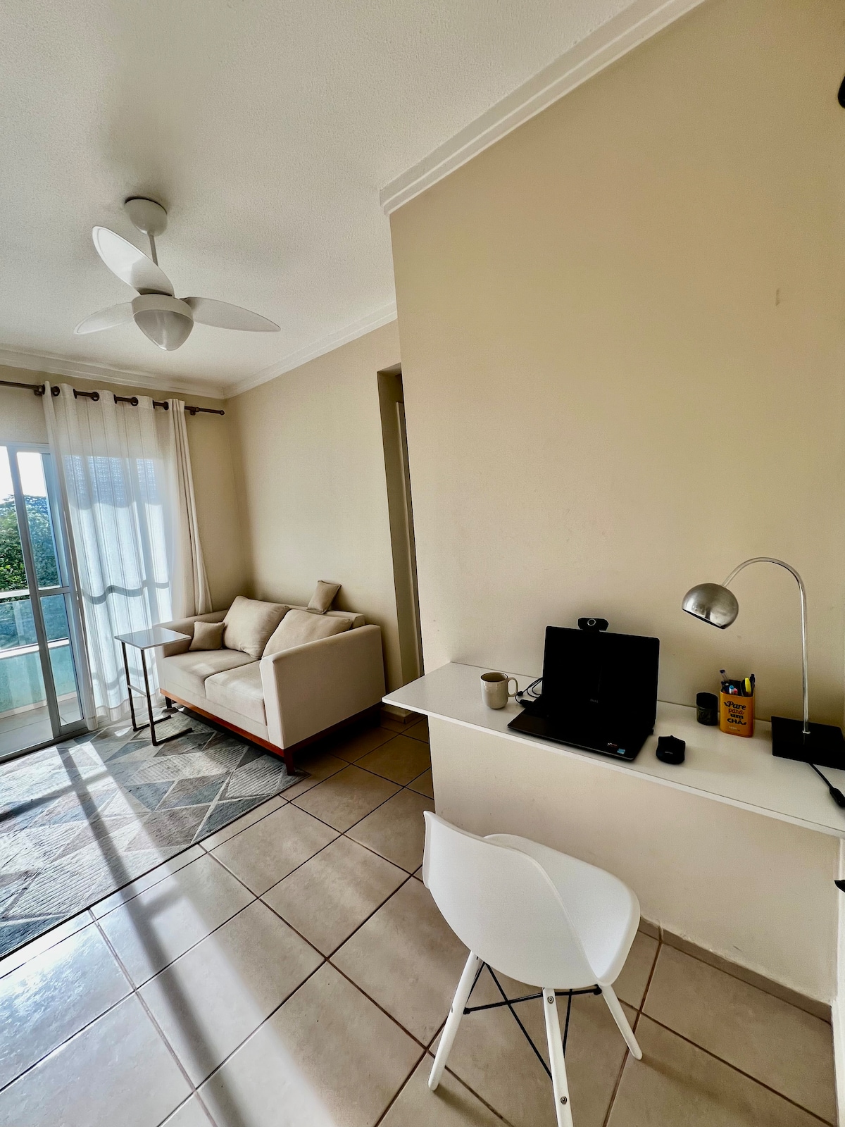 A light-filled corner features a comfortable beige sofa beside a window, offering a view of the outdoors. A tidy workspace is equipped with a laptop and organized stationery, illuminated by a sleek desk lamp. Cool tile flooring complements the soft color palette.