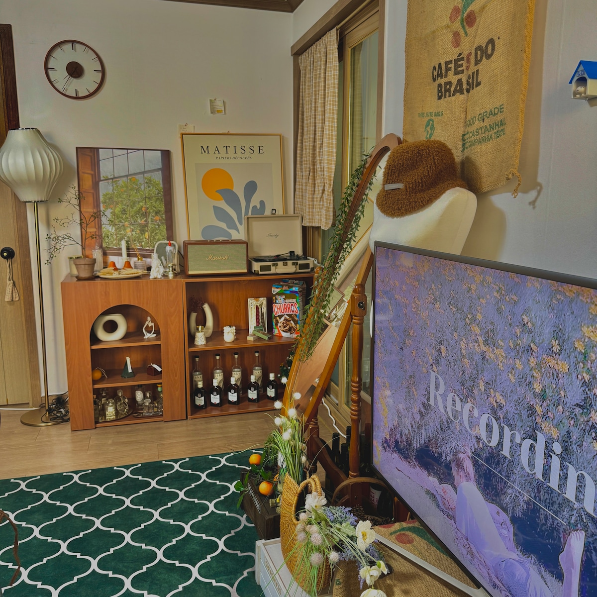 A cozy living area showcases a mix of decor, including artwork above a wooden shelf and a large television. Vintage accessories and plants are scattered throughout, while a plush rug adds comfort underfoot. Natural light filters through a nearby window, enhancing the inviting ambiance.