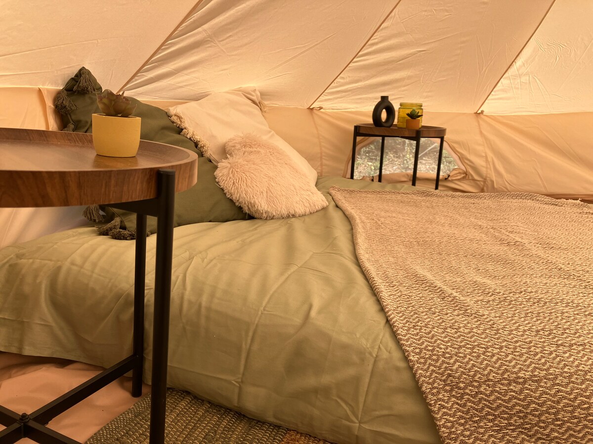 A comfortable sleeping area is featured with a spacious bed adorned with soft linens in neutral tones. Two small side tables are positioned nearby, one with a decorative vase. A cozy texture is added with a fluffy pillow and a woven throw, creating an inviting space.