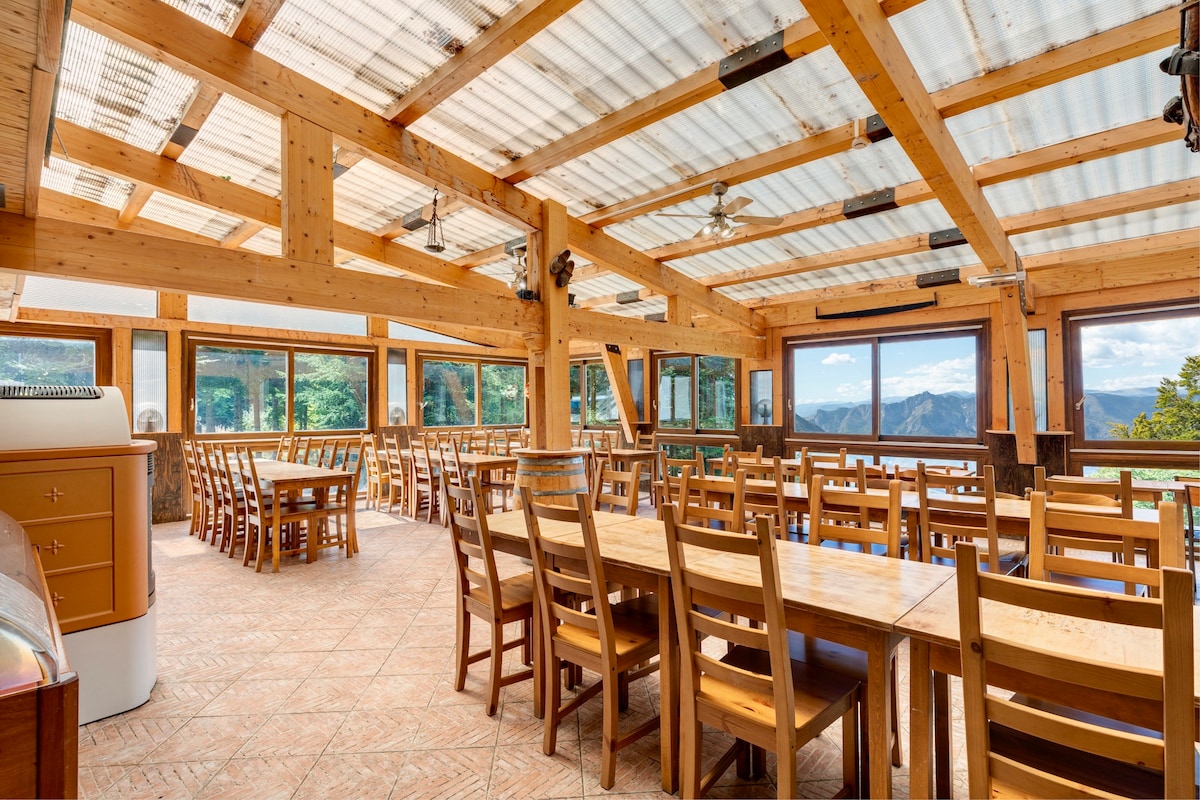 A spacious communal dining area features large windows that offer panoramic views of the surrounding mountains. Wooden tables and chairs are arranged to accommodate guests, with a warm design complemented by wooden beams and a light-colored ceiling. Ceiling fans provide comfort throughout the space.