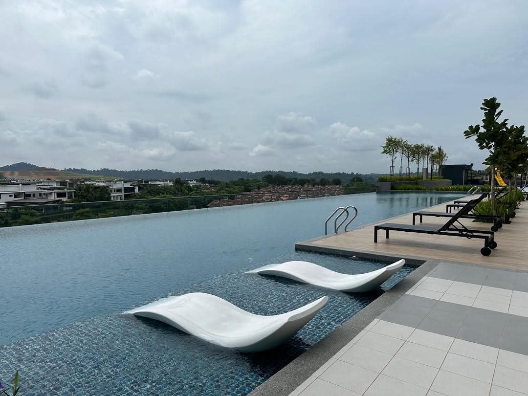 A serene infinity pool is presented, featuring two lounge chairs positioned at the water's edge. Lush greenery and a distant scenic view complement the tranquil setting. The pool surface reflects the sky, while a spacious deck area with additional lounge chairs completes the environment.